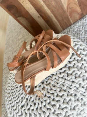Bruna kilklackade sandaler från Anna Field - Säljer ett par snygga bruna sandaler från Anna Field med kilklack i flätad jute och remmar i konstläder. Sandalerna har öppen tå och justerbart spänne runt ankeln. Perfekta för sommaren och ger en trendig look till din outfit.