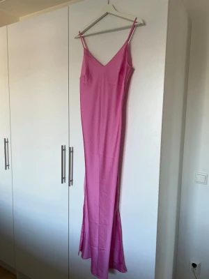 Rosa långklänning med smala axelband - Säljer en elegant rosa långklänning med smala axelband och v-ringning. Klänningen har en figurnära passform och är tillverkad i ett mjukt, slätt material som faller snyggt längs kroppen. Perfekt för dig som vill ha en stilren och trendig look.