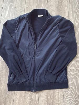 John henric cardigan  - Snygg marinblå ziptröja med stickade ärmar och ribbade muddar. Framsidan har ett slätt tyg medan ärmar och rygg är i mjuk stickad kvalitet. Hög krage och hel dragkedja framtill ger en sportig look. Priset är ej hugget i sten, skriv vid minsta fundering. Passar även S. Ända defekten på cardiganen är på dragkedjan.