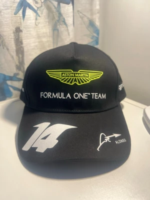 Aston Martin F1 Alonso keps  - Keps i Aston Martin Formula One Team-stil med Fernando Alonso #14. Svart med neongrön Aston Martin-logga samt BOSS, Cognizant och Aramco branding.  Bra skick. Ren och utan tydliga skador.  Justerbar storlek baktill. Perfekt för F1-fans eller till vardags.