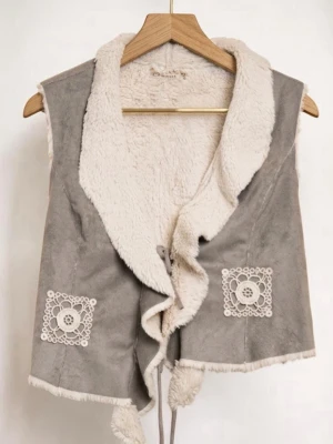 Boho Shearling-väst, fodrad  - Snygg och följsam väst med mjukt fuskpälsfoder och bred krage. Västen har dekorativa vita broderier framtill och stenar i blommönster på ryggen. Knytband framtill. Perfekt för lager på lager och trendig stil. Storlek 40 men passar 36–40. 