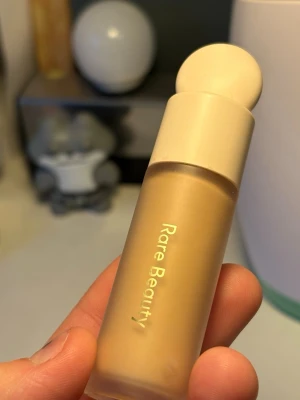 Rare Beauty Concealer 200C - Rare Beauty concealer i nyansen 200C. Kommer i en stilren, matt beige tub med rund applikator och genomskinligt fönster som visar färgen. Innehåller 7,5 ml och är perfekt för att täcka mörka ringar och ojämnheter. Lätt att applicera och smälter fint in i huden.