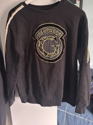 Svart sweatshirt med guldmönster - Cool svart sweatshirt med stort broderat G-märke i guld på bröstet och vita samt guldiga ränder längs axlarna. Tröjan har rund halsringning, ribbade muddar och är tillverkad i mjukt bomullsmaterial. Perfekt för en trendig och avslappnad stil.