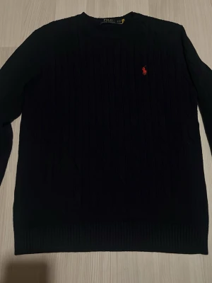 Mörkblå stickad tröja Polo Ralph Lauren - Snygg mörkblå stickad tröja från Polo Ralph Lauren med klassisk röd logga broderad på bröstet. Tröjan har rund halsringning, ribbade muddar och ett diskret kabelstickat mönster. Perfekt för dig som gillar stilrena och tidlösa plagg.