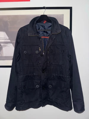 Mörkblå J.Lindberg field jacket  - Säljer en mörkblå skepparkavaj med fyra stora fickor framtill, dragkedja och knappar. Jackan har hög krage och är tillverkad i ett slitstarkt material, troligen bomull eller polyester. Perfekt för dig som gillar klassisk och funktionell stil. Vid snabb affär kan jag gå ner en hundring.