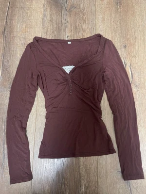 Brun långärmad topp med knappar - Snygg brun långärmad topp med v-ringning och dekorativa knappar framtill. Figurnära passform och liten spetsdetalj vid urringningen. Perfekt att styla med jeans eller kjol.