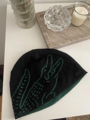 Vändbar mössa från Lacoste - Säljer en vändbar mössa från Lacoste med ikonisk krokodil på båda sidor. Ena sidan är svart med grön krokodil och den andra är grön med svart krokodil. Mössan är stickad i mjukt material och har en avslappnad passform. Perfekt för dig som vill ha två stilar i en.