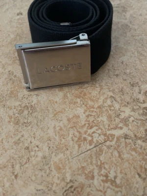 Svart Lacoste bälte 100cm - Snyggt svart tygbälte från Lacoste med silverfärgat spänne där loggan är ingraverad. Bältet har den klassiska gröna krokodilen broderad i ena änden och passar perfekt till jeans eller chinos. Bältet är okej skick fast lådan är lite trasig i formen 