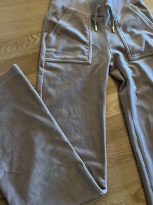 Beige mjukisbyxor Juicy Couture - Säljer ett par beige mjukisbyxor från Juicy Couture med broderad logga och guldfärgade detaljer på snöret. Byxorna har raka ben, resår i midjan och stora fickor framtill. Tillverkade i mjuk velour som känns riktigt skön mot huden.