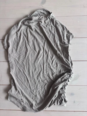 Grå kortärmad topp med rynk - Säljer en enkel grå topp i mjuk bomull med korta ärmar och snygg rynkdetalj på ena sidan. Perfekt till jeans eller kjol för en avslappnad look.