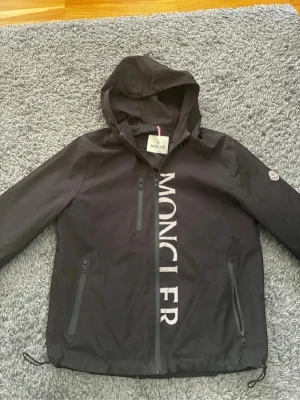 Svart Moncler vindjacka med huva - Svart vindjacka från Moncler med stor vit logga längs dragkedjan framtill. Jackan har tre dragkedjeförsedda fickor, justerbar nederkant och en fast huva. Materialet är lätt och vattenavvisande, perfekt för blåsiga dagar. Klassisk Moncler-patch på ärmen.