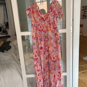 Blommig långklänning med puffärm - Färgglad långklänning med blommigt mönster i rosa, rött, lila och vitt. Klänningen har puffärmar och en söt, romantisk look med volangdetaljer vid axlarna. Perfekt för sommarens alla tillfällen.