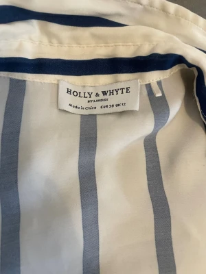 Randig skjorta från Holly & Whyte - Snygg skjorta från Holly & Whyte by Lindex i vitt med blå vertikala ränder. Skjortan har klassisk krage och långa ärmar. Perfekt för en stilren och avslappnad look. Tillverkad i ett mjukt och lätt material som känns skönt mot huden.