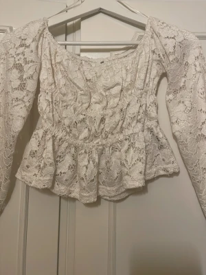 Vit spetsblus med volang från H&M - Superfin vit blus i spets från H&M med volang nertill och långärmade puffärmar. Blusen har en romantisk känsla med sitt genombrutna spetsmönster och bred urringning. Perfekt att styla med jeans eller kjol för en trendig look.