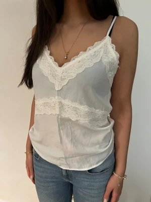 Vit spetslinnetopp med smala band - Säljer en vit linnetopp med tunna axelband och v-ringning. Toppen har fina spetsdetaljer längs urringningen och tvärs över bröstet. Materialet känns lätt och svalt, perfekt för varma dagar. Enkel att styla med jeans eller kjol.