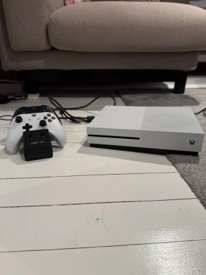 Xbox One S vit spelkonsol med handkontroll - Säljer en Xbox One S i snygg vit design med tillhörande vit och svart trådlösa handkontroller med laddstation . Konsolen har en stilren, rektangulär form och är tillverkad i plast. En av dom behöver att man byter batteri på (har för mig att det är den vita). Det har också lite märke på kontrollerna.Men det är perfekt för gaming och streaming.