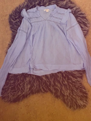 Ljusblå blus med volanger - Söt ljusblå blus med volangdetaljer och spetsinslag vid axlar och ärmar. Blusen har v-ringning och är tillverkad i ett tunt, luftigt tyg.