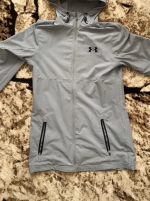 Grå vindjacka från Under Armour - Snygg grå vindjacka från Under Armour med huva och hel dragkedja framtill. Jackan har två svarta dragkedjefickor och en svart logga på bröstet. Tillverkad i lätt och vindtätt material, perfekt för blåsiga dagar.