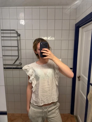 Vit blus med volangärmar - Söt vit blus med tunna vertikala ränder och volangdetaljer längs ärmarna. Blusen har rund hals med spetskant och är tillverkad i ett lätt, luftigt tyg. Perfekt för en trendig och feminin look.