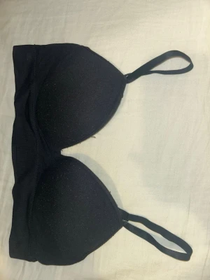 Svart bandeau-bh med vaddering - En svart bandeau-bh med tunna, justerbara axelband och vadderade kupor. Bh:n har en slät design utan synliga sömmar eller dekorationer och är tillverkad i ett stretchigt material för extra komfort.