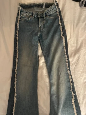 Blå bootcut jeans med fransade kanter - Säljer ett par blå bootcut jeans med råa, fransade kanter längs sidorna och vid linningen. Jeansen har klassisk femficksdesign och är tillverkade i denim med lätt tvättad look. Perfekta för dig som vill ha en unik och trendig stil.