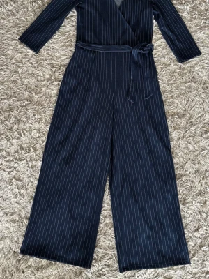 Mörkblå randig jumpsuit Bershka - Snygg mörkblå jumpsuit från Bershka med vita tunna ränder. Modellen har långa ärmar, omlottopp och ett matchande knytband i midjan. Vida ben ger en avslappnad men trendig look. Perfekt för dig som gillar stilrena plagg med lite edge.