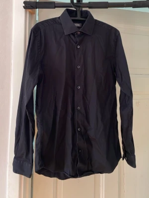 Svart skjorta från Dressmann - Stilren svart skjorta med klassisk krage och knappar framtill. Skjortan har lång ärm med manschett och är tillverkad i mjuk bomull. Endast använd ett fåtal gånger, storlek M 39/40 Slim fit 