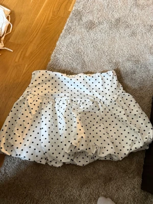 Vit prickig kortkjol med volang - Söt vit kortkjol med svarta prickar och volangkant. Kjolen har en ballongliknande form och bred midja som ger en trendig siluett. Perfekt för dig som gillar lekfulla detaljer och klassiskt mönster.