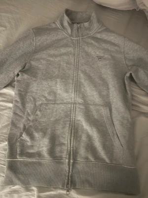 Grå ziptröja från GANT - Snygg grå ziptröja från GANT med hög krage och två fickor framtill. Tröjan har GANT-logga broderad på bröstet och ribbade muddar vid ärmslut och nederkant. Insidan av kragen har en stilren randig detalj. Perfekt för en avslappnad men stilren look.