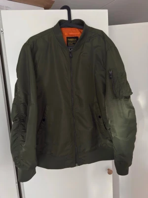 Olivgrön bomberjacka  - Snygg olivgrön bomberjacka  med orange innerfoder. Klassisk modell med ribbade muddar, dragkedja framtill och fickor på sidorna samt en ärmficka med dragkedja. Perfekt för dig som vill ha en stilren och trendig jacka.
