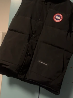 Svart dunväst från Canada Goose - Snygg svart dunväst från Canada Goose med klassisk logotyp på bröstet och stora praktiska fickor framtill. Västen har tryckknappar och är tillverkad i slitstarkt material med värmande dunfyllning. Perfekt för lager-på-lager och streetwear.