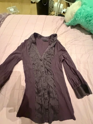 Lila blus med volang och knappar - Snygg lila blus från H&M med långa ärmar och breda manschetter. Blusen har volangdetaljer och knappar framtill samt en djup V-ringning. Perfekt för att lyfta din outfit med en unik touch.