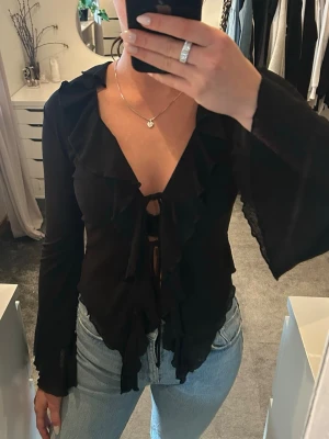 Svart volangblus med knytning - Trendig svart blus med långa ärmar och volangdetaljer längs urringningen och framtill. Blusen har djup v-ringning och knytning framtill som ger en cool och feminin look. 