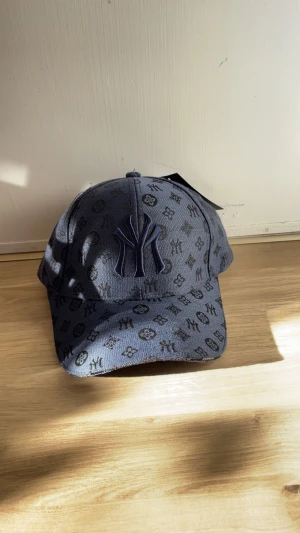 Mörkblå  keps med monogram - Snygg mörkblå keps med NY-logga framtill och all-over monogrammönster. Kepsen har böjd skärm och justerbar rem bak. Tillverkad i ett slitstarkt tygmaterial som ger en cool streetwear-vibe.