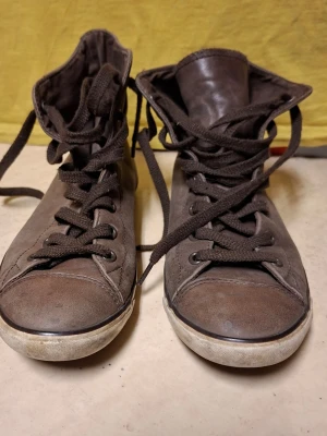 Bruna höga sneakers i läder, converse chuck taylor all star - Säljer ett par bruna höga converse med klassisk snörning och slitstark gummisula. Skorna är tillverkade i läder och har en enkel, stilren design som passar till det mesta. Perfekta för dig som gillar en clean och tidlös look.