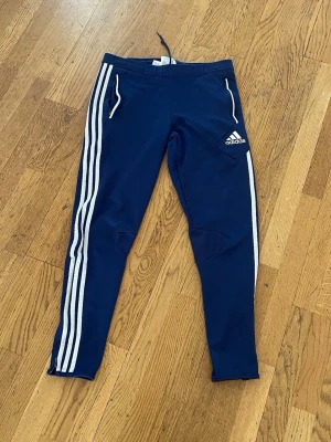 Blå Adidas träningsbyxor med ränder - Mörkblå träningsbyxor från Adidas med klassiska vita ränder längs sidorna och vit Adidas-logga på vänster lår. Byxorna har dragkedjor vid bensluten och vita dragkedjor på fickorna. Tillverkade i ett stretchigt funktionsmaterial, perfekta för träning.