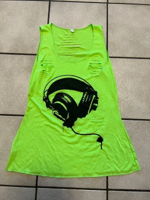 Neongrön linnetopp med hörlursmotiv storlek L - Cool neongrön linnetopp med svart tryck av stora hörlurar på framsidan. Toppen har flera slitna detaljer framtill och är ärmlös med rund halsringning. Perfekt för dig som gillar streetstyle och vill sticka ut.