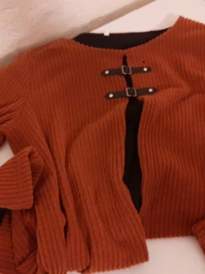 Rostbrun stickad kofta med spännen - Säljer en ribbstickad kofta i rostbrunt med två dekorativa svarta spännen framtill. Koftan har rund halsringning och svarta detaljer på insidan. Perfekt för dig som gillar en unik och trendig look.