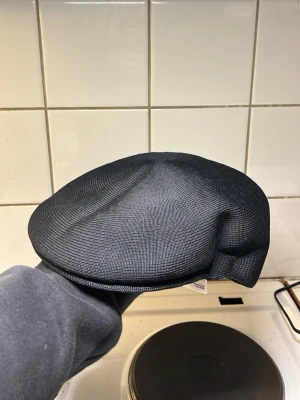 Svart Kangol x J.Lindeberg keps - Snygg svart keps i klassisk flat cap-modell från samarbetet mellan Kangol och J.Lindeberg. Kepsen har en stilren struktur i tyg och en broderad logga bak. Perfekt accessoar för en trendig och avslappnad look.