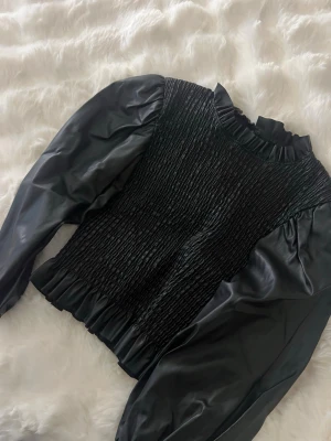 Svart topp med puffärmar i skinnimitation - Trendig svart topp med puffiga långärmar i skinnimitation och smockad kropp. Hög krage med volangdetalj och elastisk midja för en snygg passform. Perfekt för dig som vill sticka ut med en edgy look.