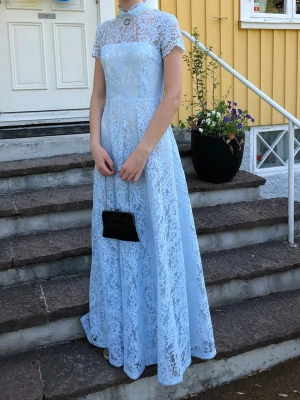 Ida Sjöstedt Ljusblå lång spetsklänning - Elegant ljusblå klänning i spets med hög hals och korta ärmar. Klänningen har en figurnära överdel och ett långt, svepande A-linjeformat kjolparti. En unik klänning till bal. Endast buren en gång. Från Ida Sjöstedt, säljs inte längre. Nypris 9000 kr