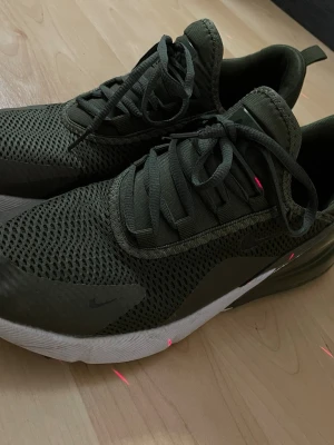 Nike Air Max 270 olivgröna sneakers - Nike Air Max 270 sneakers i olivgrönt med mesh-överdel och tydlig AIR-dämpning i hälen. Skorna har svarta detaljer, snörning och en vit sula som kontrast. Perfekta för dig som gillar sportig stil och vill ha bekväma sneakers med modern design.