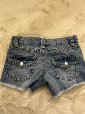 Blå jeansshorts med fransig kant - Snygga blå jeansshorts med fransig kant och slitna detaljer. Shortsen har fem fickor, bälteshällor och knappar i silverfärg. Perfekta för varma dagar och ger en avslappnad look. Tror det är storlek 152/158