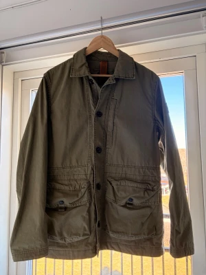 Olivgrön overshirt från BOSS - Stilren olivgrön overshirt från BOSS i bomull. Klassisk krage, stora framfickor med knappdetaljer och knappstängning framtill. Patch med BOSS-logga på ärmen ger en snygg touch. Perfekt lager-på-lager-plagg för en avslappnad look. Säljer nu min snygga overshirt ifrån BOSS! Säljer den eftersom den var lite stor för mig. Overshirten är använd 1 gång så är som nyskick.Tveka inte på att höra dig och ställa frågor om du har några🤩😁