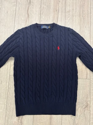 Mörkblå kabelstickad tröja Polo Ralph Lauren - Kabelstickad mörkblå tröja från Polo Ralph Lauren med rund halsringning och röd broderad logga på bröstet. Knappast använd. Pris kan diskuteras! 