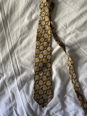 Vintage slips från Yellow Springs - Säljer en snygg slips från Yellow Springs med geometriskt mönster i gult, beige och mörkblått. Slipsen har en klassisk form och är tillverkad i ett glansigt tyg som ger en elegant känsla. Perfekt för dig som vill sticka ut med accessoarerna.