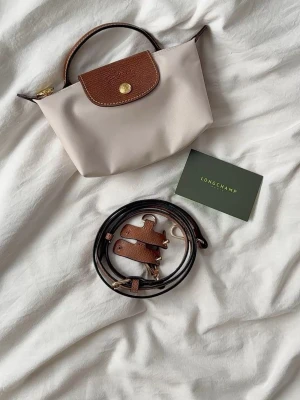 Beige och brun handväska - Beige handväska med bruna läderdetaljer och guldfärgade metalldetaljer. Väskan har ett klassiskt lock med tryckknapp och präglad logga samt ett kort handtag i läder. Axelrem tillkommer och prislappar tillkommer.💕