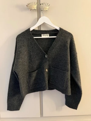 Mörkgrå kofta från AWARE - Mysig mörkgrå kofta från AWARE med två stora fickor framtill och v-ringad hals. Koftan har en avslappnad, boxig passform, ribbade muddar och stängs med tre knappar. Perfekt för lager på lager och enkel att styla till olika outfits. Säljer denna kofta då jag aldrig använt den. Pris kan diskuteras! 