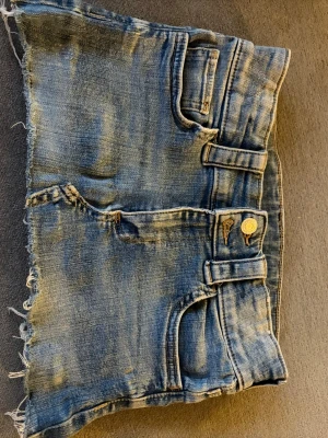 Blå jeanskjol med rå kant - Cool kort jeanskjol i blå denim med slitna detaljer och rå, fransig kant. Kjolen har klassiska fickor fram och bak samt bälteshällor och knappstängning. Perfekt för dig som gillar en avslappnad och trendig look.