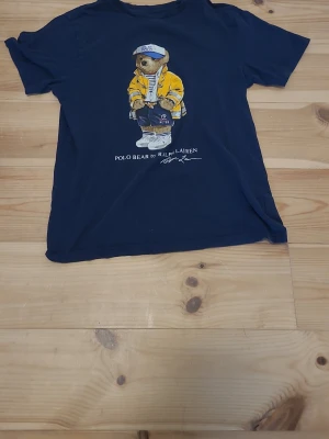 Mörkblå Polo Bear t-shirt Ralph Lauren - Säljer en mörkblå t-shirt från Ralph Lauren med den ikoniska Polo Bear-trycket på bröstet. T-shirten har rund hals och korta ärmar. Tillverkad i mjuk bomull som känns skön mot huden. Perfekt för dig som gillar streetwear och klassiska prints.